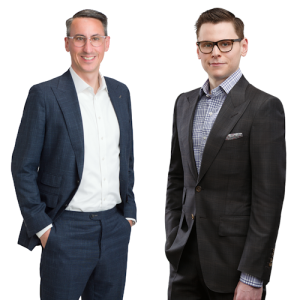 A portrait of two men from Scott Telecom Services. From left to right: Ryan Scott, President, wearing glasses and a dark suit with a white shirt, smiling with his hands in his pockets; and Hunter Scott, Executive Vice President, wearing glasses and a dark suit with a checkered shirt, smiling with his hands in his pockets. Both are standing against a white background, conveying a sense of professionalism and confidence.